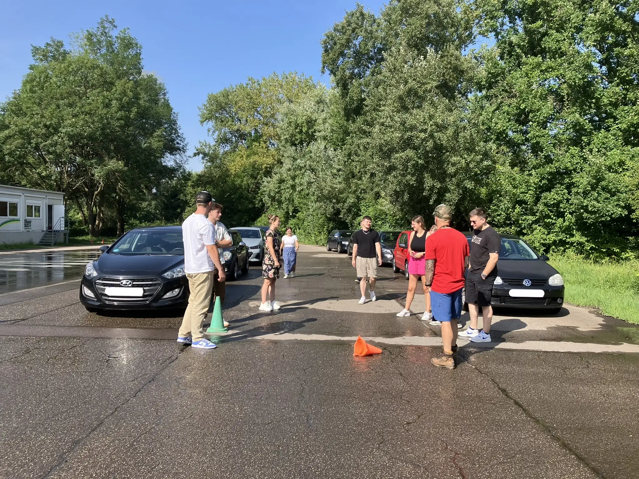 bardusch-Azubis stehen mit dem Trainer um ein Fahrzeug und besprechen die Fahr&uuml;bung zwischen Pylonen auf nasser Strecke