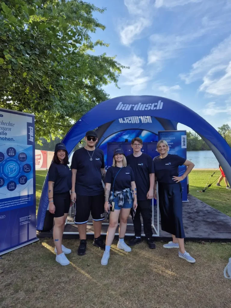 Azubis von bardusch stehen in Teamkleidung vor dem blau-wei&szlig;en bardusch-Pavillon auf dem SunnyLake Festival.