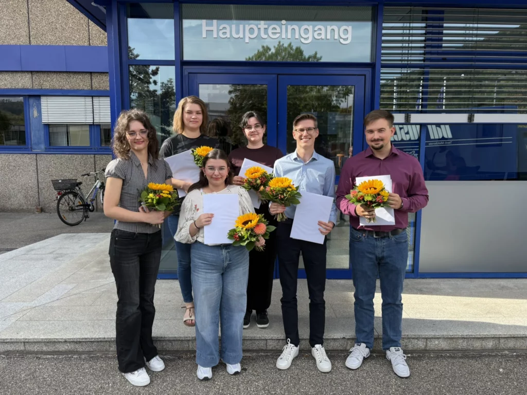 Absolventinnen und Absolventen mit Blumensträußen und Zeugnissen vor dem Haupteingang von bardusch