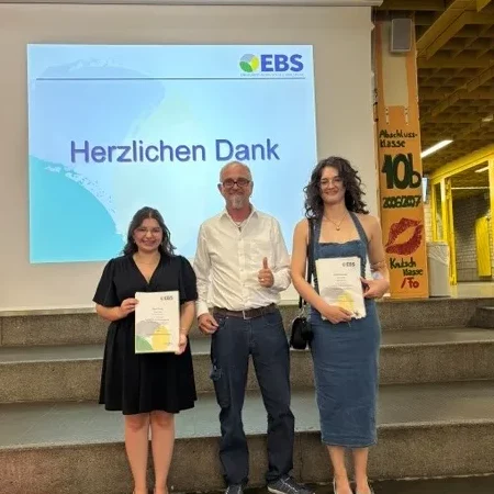 Drei Personen bei einer feierlichen Ehrung mit Urkunden und Blumen auf einer Bühne